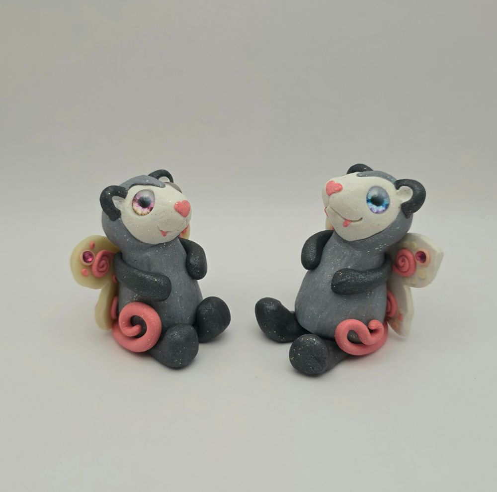 Two little fairy opossums  sitting facing each other with swirly tails, heart shaped noses, wings, colorful eyes, and pink fangs.