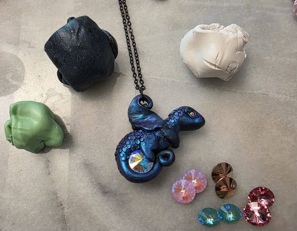 A shiny blue dragon pendant with its tail wrapped around a rivoli crystal, surrounded by more crystals and lumps of clay
