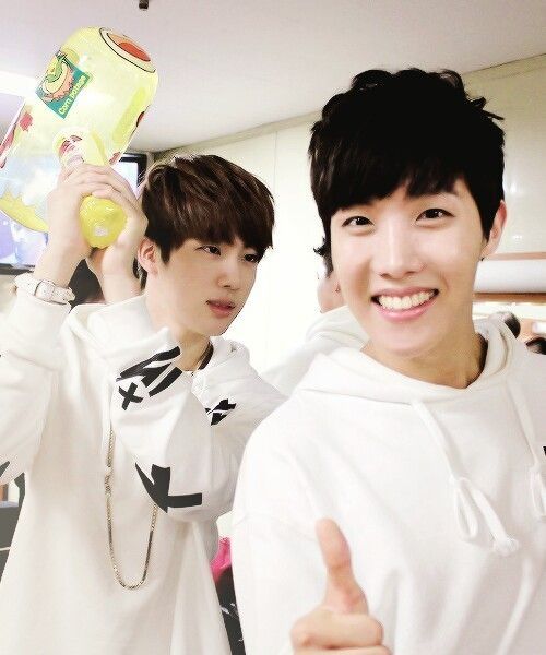 Young hobi and jin from a bangtan bomb. Jin is holding an inflated hammer(?) And is ready to bonk hobi. Hobi is smiling at the camera with a thumbs-up sign. They are both in white outfits.