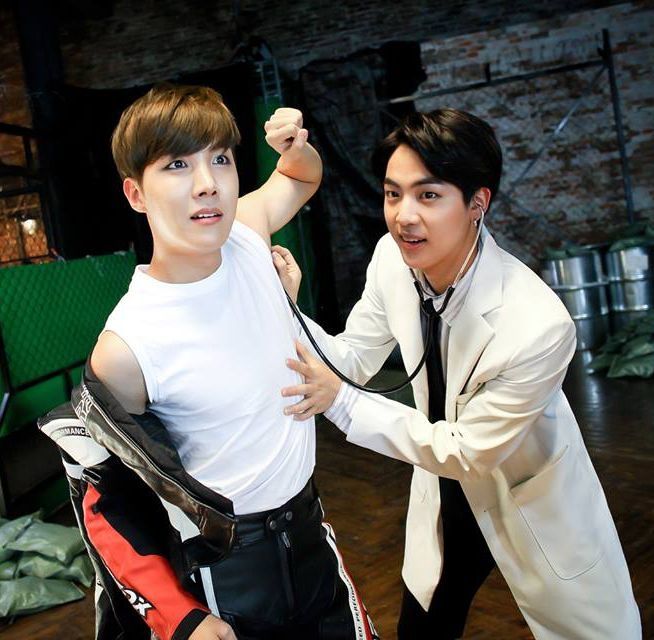 Jin and hobi on the "dope" set. Jin in his doctor outfit has placed one end of his stethoscope to hobi's armpit. Hobi is standing in a white singlet, his racer jacket hanging on the other arm and he has a funny expression on his face.