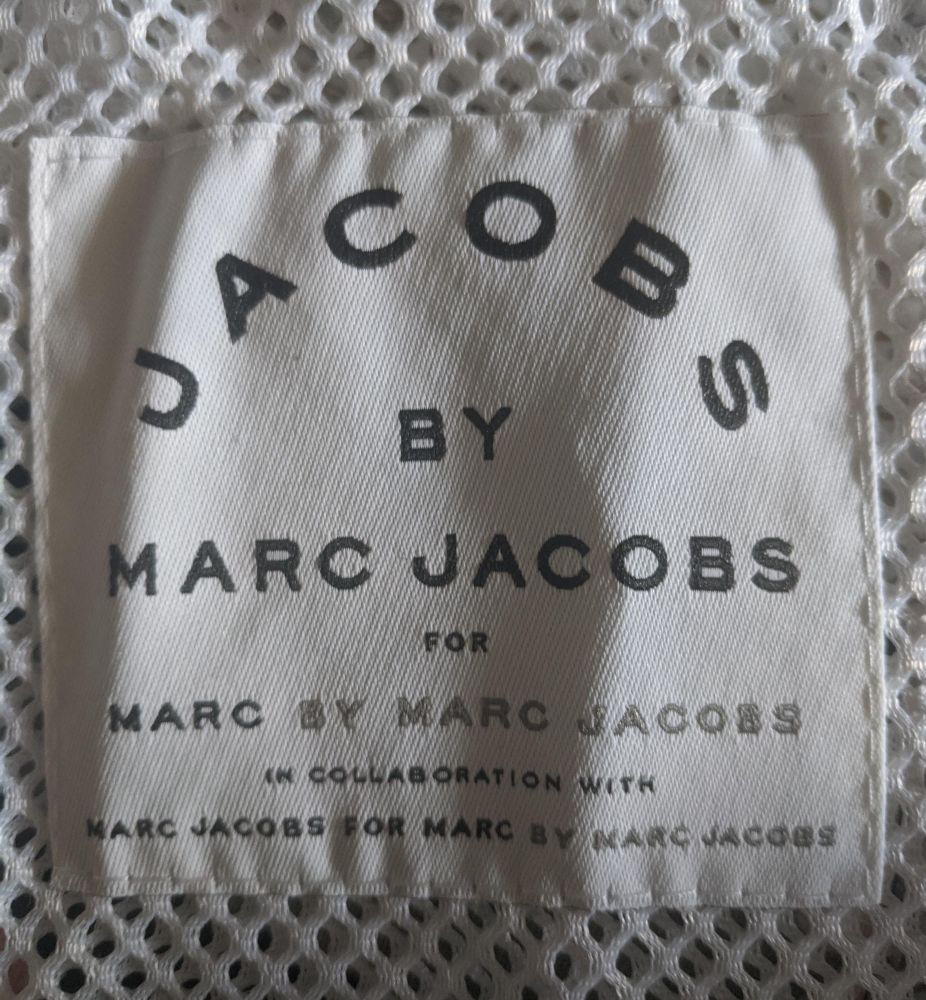 A cream-colored fabric clothing label with black text reading “JACOBS BY MARC JACOBS FOR MARC BY MARC JACOBS IN COLLABORATION WITH MARC JACOBS FOR MARC BY MARC JACOBS” arranged in a centered, stacked layout. The label sits on white crocheted or mesh lace fabric with an open honeycomb pattern.​​​​​​​​​​​​​​​​