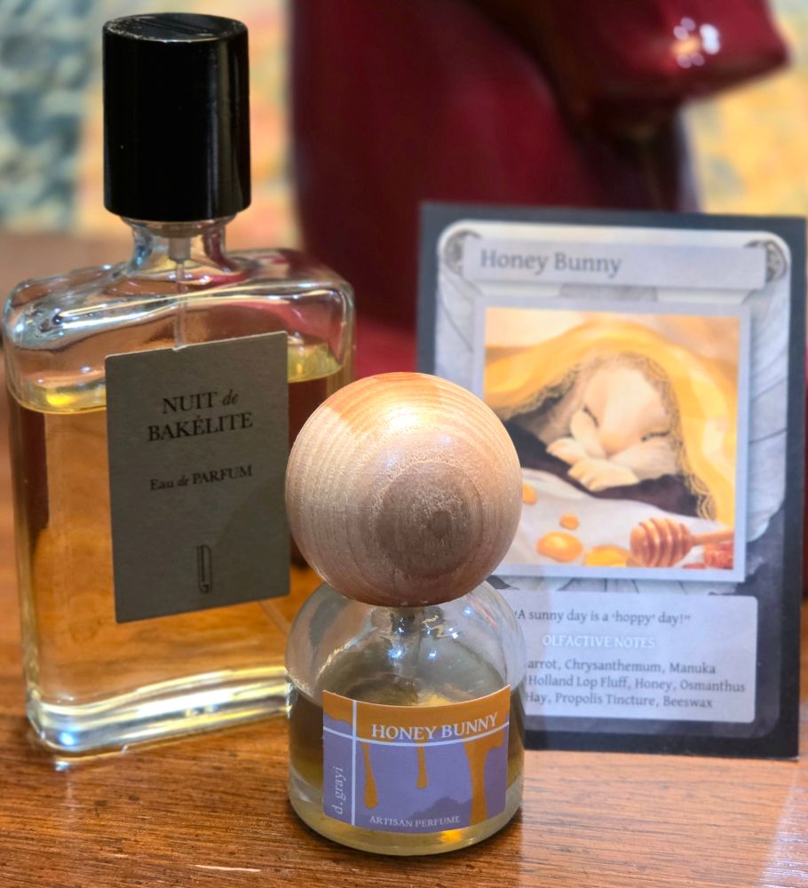 A bottle of Nuit de Bakélite that is approximately 75% full next to a bottle of Honey Bunny that is about half full. In the background is the collector's card that accompanies Honey Bunny, which shows an illustration of a white bunny sleeping under a yellow blanket with a honey drizzler and a list of olfactive notes.