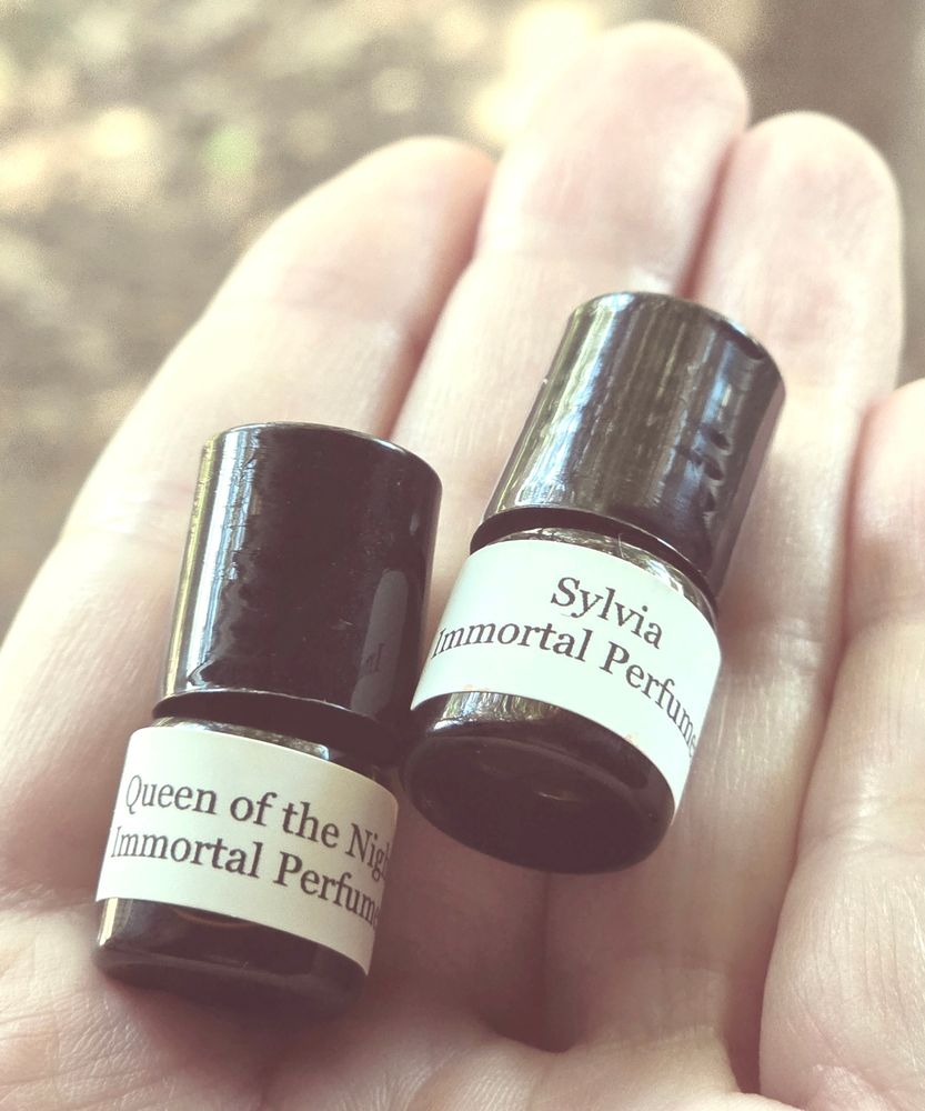 Two mini perfume oil roller vials made of brown glass with black caps laying in the palm of my hand. Perfume names are Queen of the Night and Sylvia.