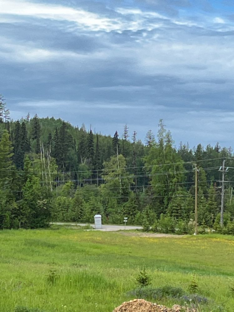 Grey portable toilet at the edge of a road and hayfield, with a forested hill and cloudy sky in the background.
