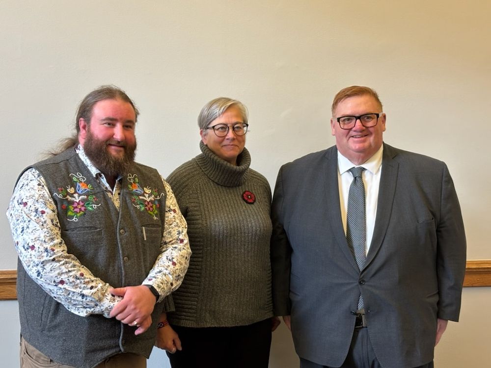 Three people standing indoors against a plain wall, dressed in formal and semi-formal attire, posing for a group photo.