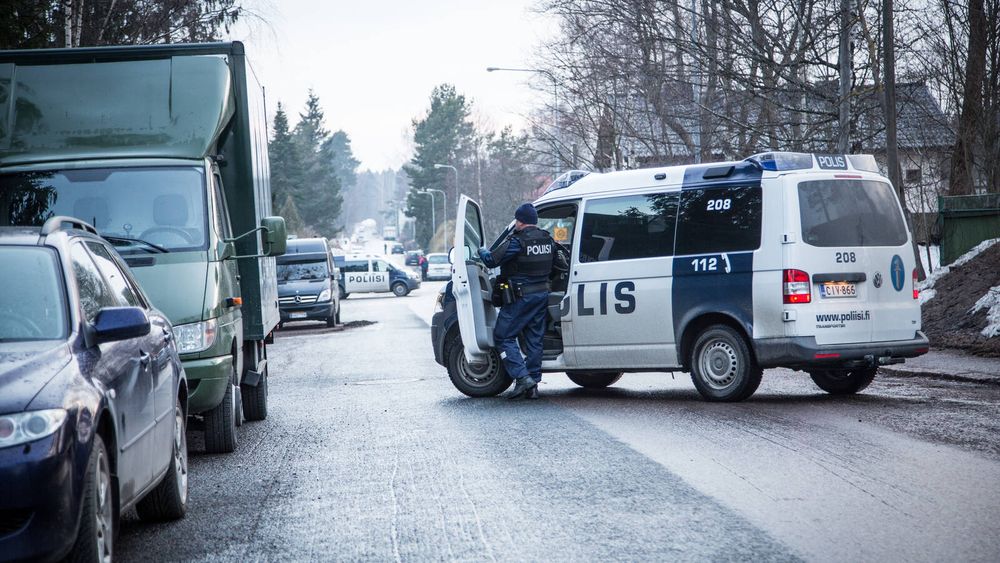 Valokuvassa poliisi poliisiauton vieressä. Auto on pysähtynyt asuinkadulle. Taustalla kauempana näkyy myös toinen poliisiauto.
