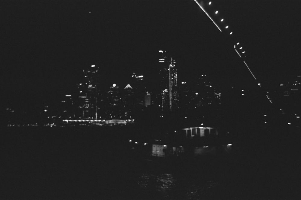 Am underexposed panchromatic cityscape of Sydney over the water, the lights of a ferry in the foreground 