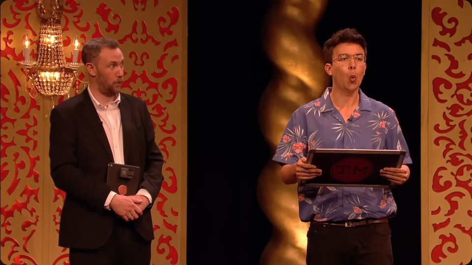 Screenshot from the live studio task in Taskmaster S7E3 of Alex Horne looking over at and Phil Wang mouthing a fact.
