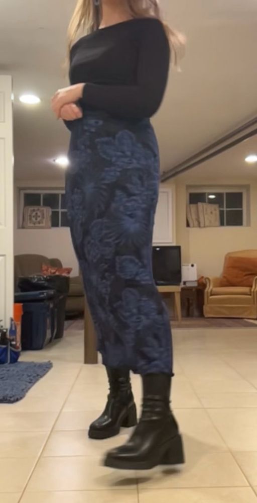 Photo of woman wearing black long sleeve with a dark blue skirt and black skinny faux leather boots. The blue skirt has swirling flower patterns on it.