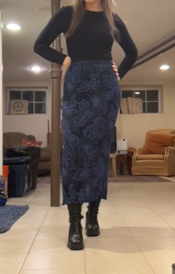 Photo of woman wearing black long sleeve with a dark blue skirt and black skinny faux leather boots. The blue skirt has swirling flower patterns on it.