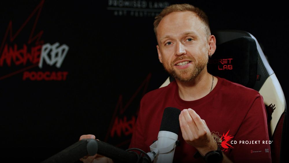 Paweł Burza sits chatting on the set of AnsweRED Podcast, looking directly towards the camera. His hand is raised, fingers pursed. The CD Projekt Red logo is printed in the bottom right corner.