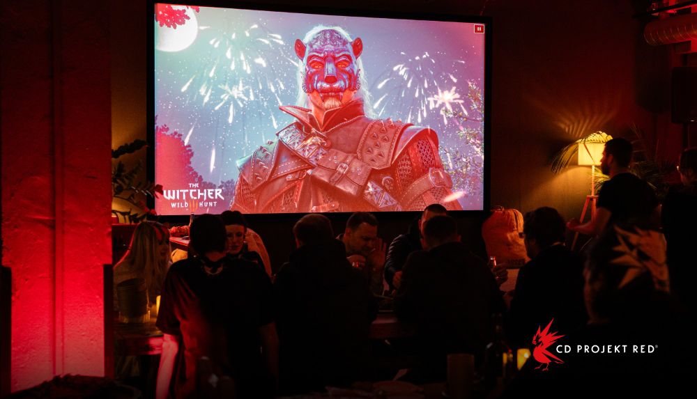 A group of CD Projekt Red employees sit at a table and talk. A large screen behind them shows Geralt of Rivia wearing a masquerade mask.