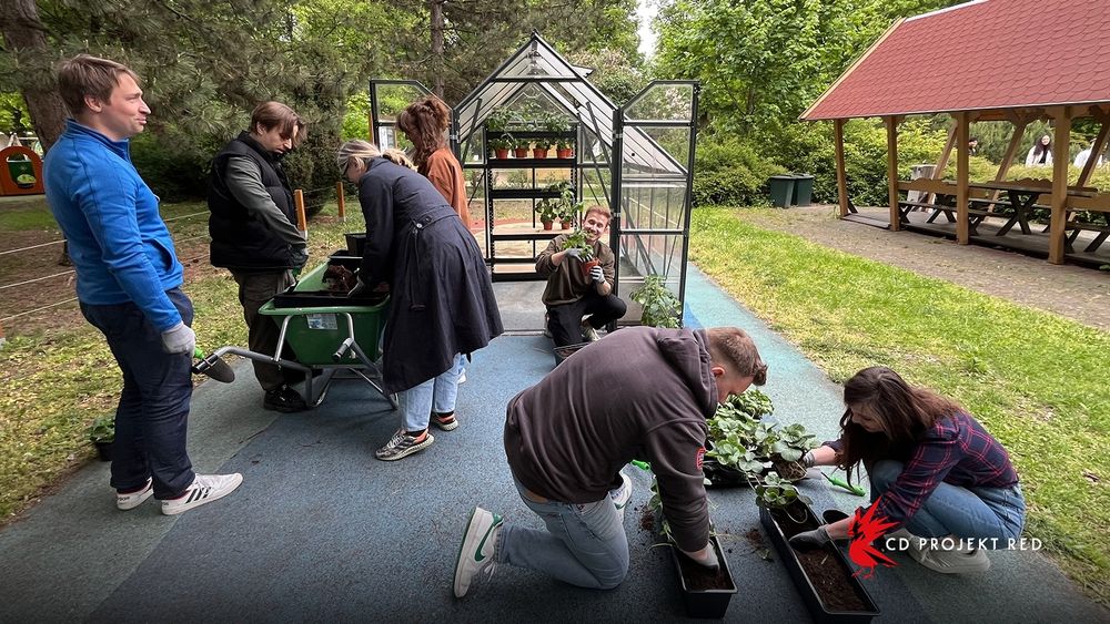 CD Projekt Red employees plant flowers and crowd around a wheelbarrow outdoors.