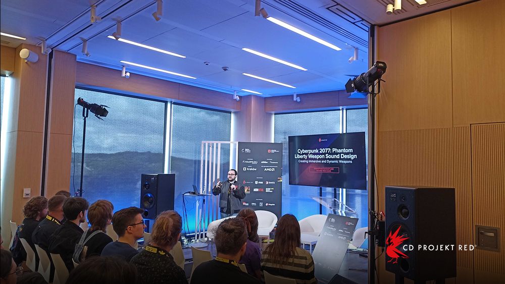 A CD Projekt Red employee addresses a seated audience. Next to him, a TV reads, “Cyberpunk 2077: Phantom Liberty Weapon Sound Design.”