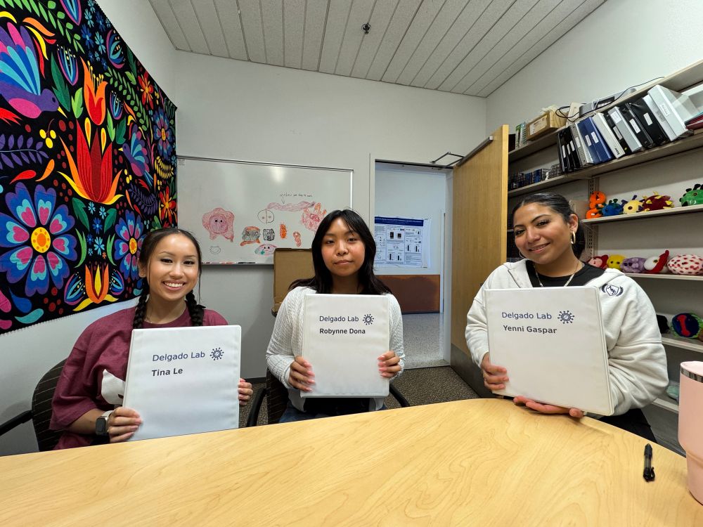 Three young women holding a binder that says “Delgado lab” in a office 