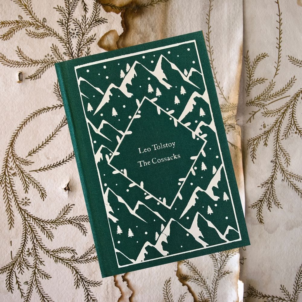 A green hardcover edition of The Cossacks by Leo Tolstoy rests on a textured background with botanical illustrations and burn marks. The book cover features a repeating white design of snow-capped mountains and pine trees, framed by a diamond-shaped border around the title.
