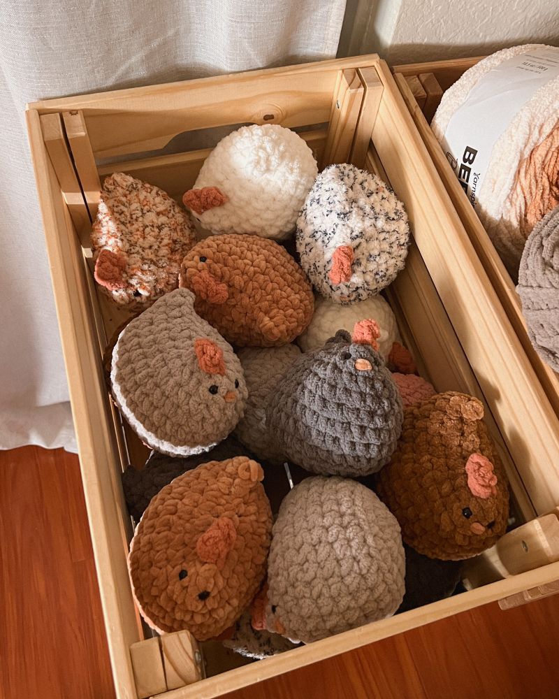 Crate full of multicolored crochet chickens. The chickens are shades of white, cream, speckled, and brown. 