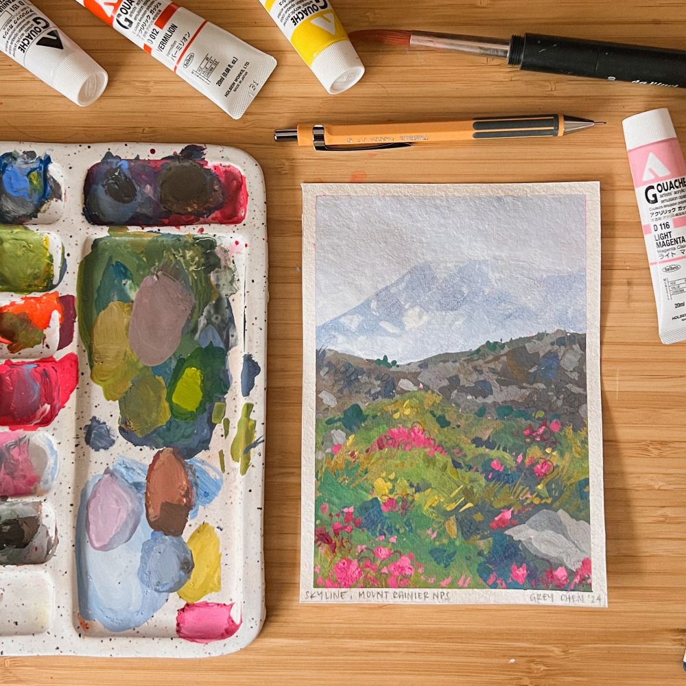 Photo of a gouache painting featuring Mount Rainier on the Skyline Trail. Foreground has a lot of vibrant pink and yellow flowers with the mountain fading into the background. Painting is on a desk next to paint tubes, pencils and brushes, and a used paint palette with the colors used to create an aesthetic composition