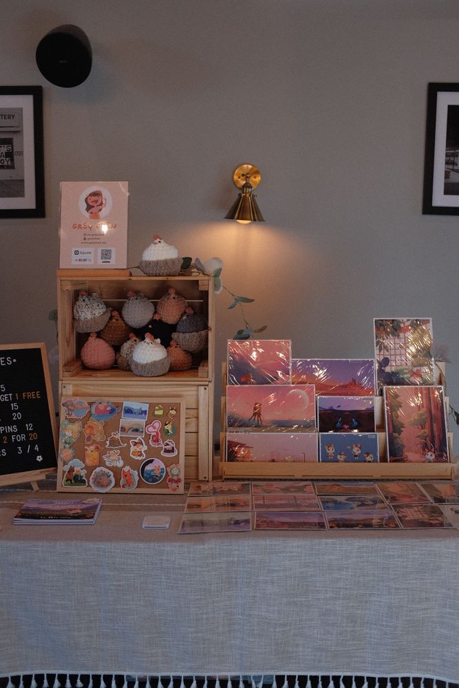 Photo of an artist’s vendor set up. On the left is a price board and crates stacked on each other full of crochet chickens. There is a sticker board display. On the right is a tiered stand of prints and postcards on the table