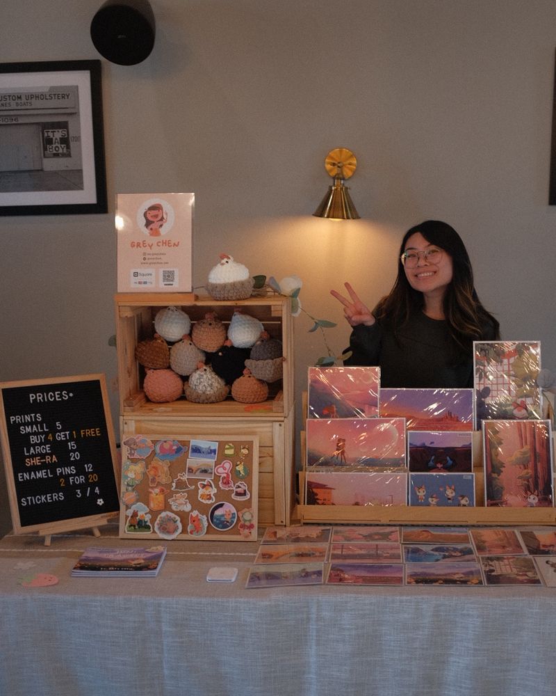 Photo of girl standing behind her market set up. The table is decorated with postcard prints of illustrations of travel destinations, stickers, art books, and crochet items. The interior is inside a cozy coffee shop. 