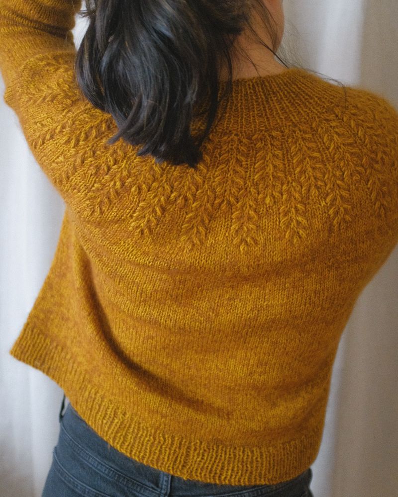 Girl modeling a hand knit sweater: the sweater is a goldenrod yellow color with brown marling, the yoke features a textured wheat grain pattern that starts at the neck and flares out down to the chest. The rest of the sweater is knit using stockinette stitch
