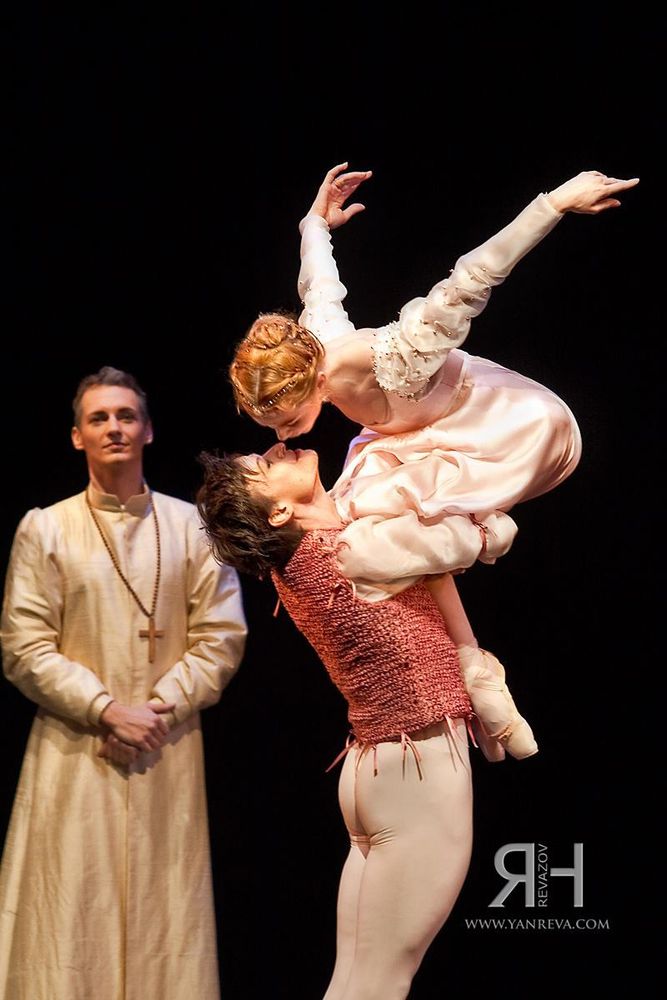 Photo from a ballet production showing a man holding up a woman possibly mid-jump, her knees pulled up to her chest and her rear ending up over his crossed arms around her calves. Her arms are stretched back and out as she leans her head toward his for a kiss. Meanwhile, another man dressed as a priest stands nearby facing the viewer, his hands clasped in front of him with a fairly neutral expression, though slightly smiling