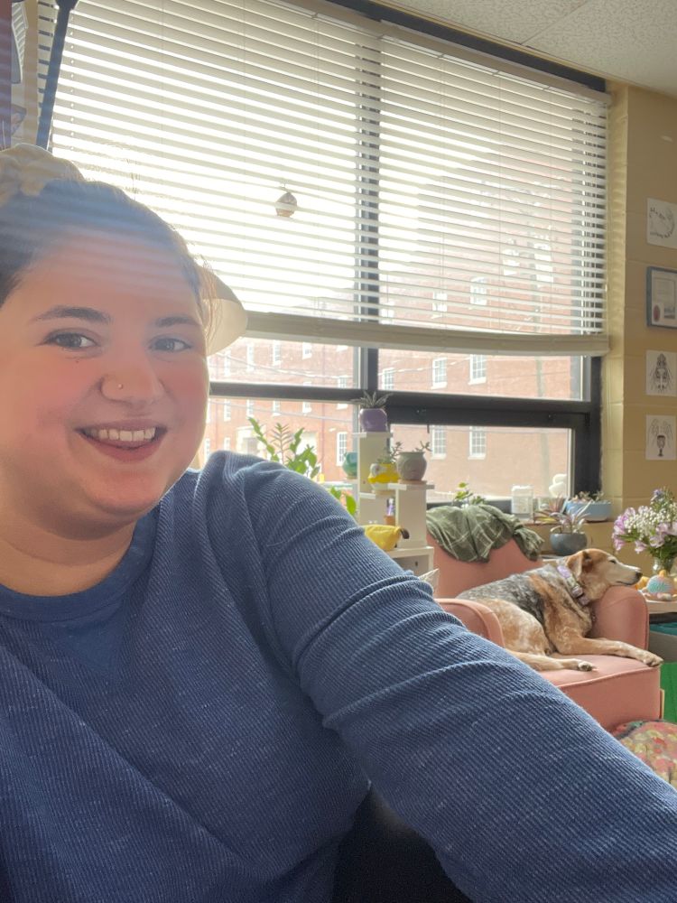 mads, smiling, with a cozy blue long sleeve shirt on. in the background is astella (a cattle dog mix) on a pink chair. there are also plants in the background and a large window