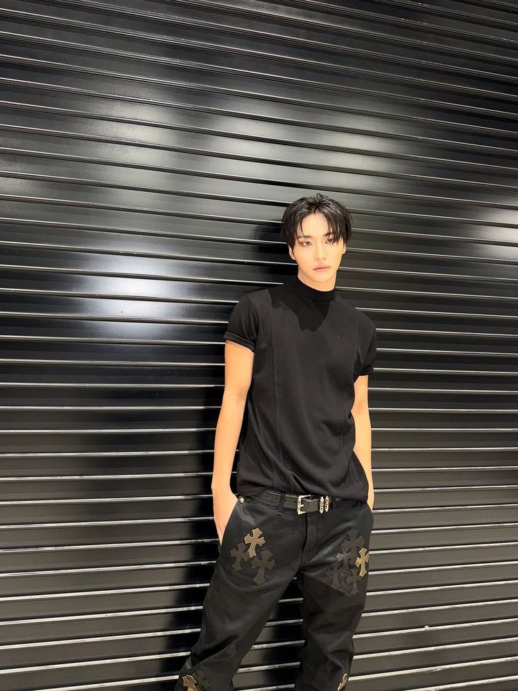 Seonghwa dressed in all black, posing in front of either a metal bat door or wall with him his hands in his jeans pockets. His jeans have crosses on them and his shirt is short sleeved.