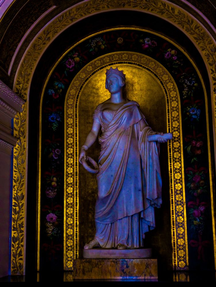 Chromatic colour edit of a statue of young queen victoria, dressed in a robe and wear her crown,  rich golds surround her in the background and a mosaic of colourful flowers. 