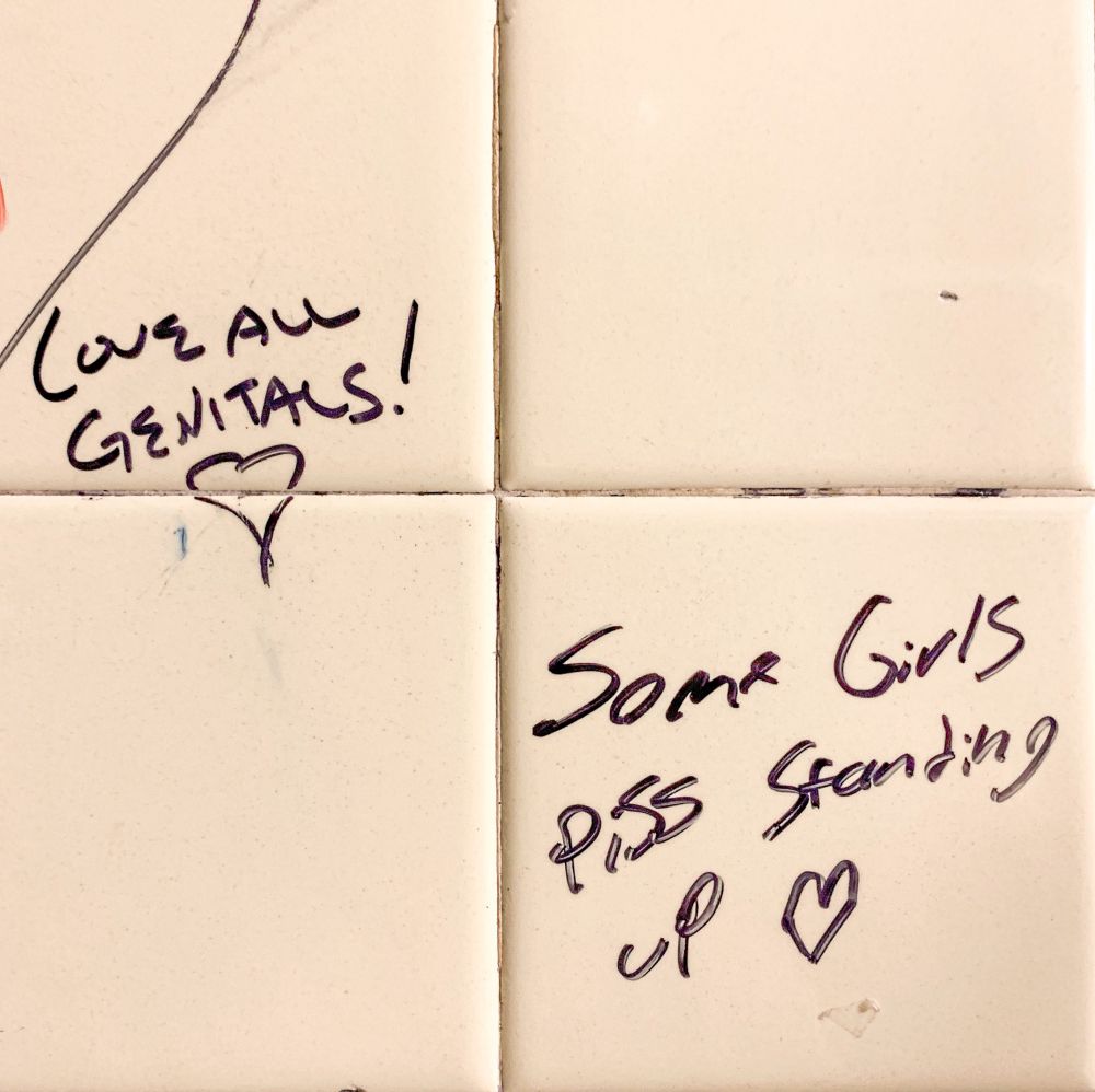 Black graffiti writing on white tile saying "Love all genitals!" and "Some girls piss standing up"