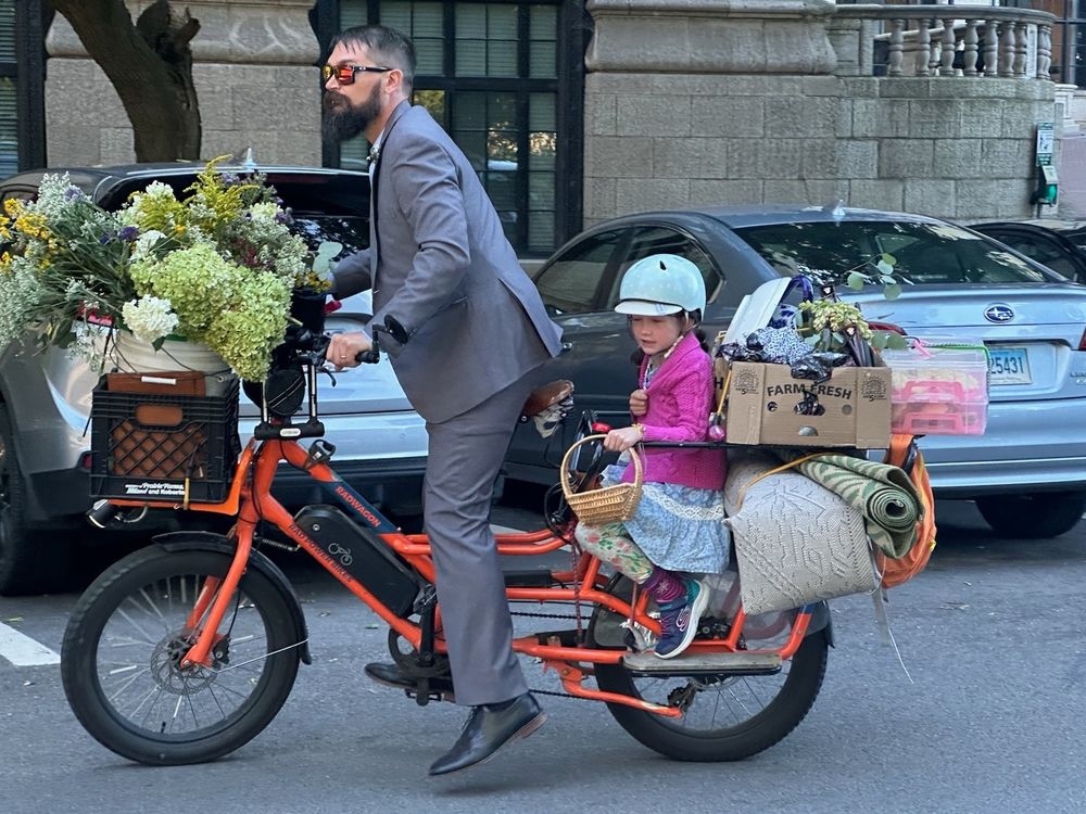 Man on cargo bike in gray suit with small child on back, hauling flowers and other wedding decoration. 