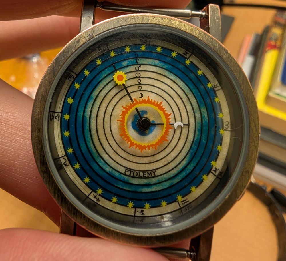 A watch depicting the Ptolemaic universe: the sun and moon are hands, the signs are hours, 7 planetary symbols turn as seconds over a ringed background symbolizing the planetary spheres.
