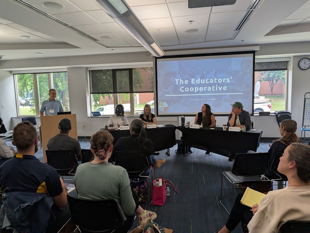 A photograph of a moderator at a podium who is next to a panel of 4 educators all before an audience of at least ~7 educators in a classroom setting.
