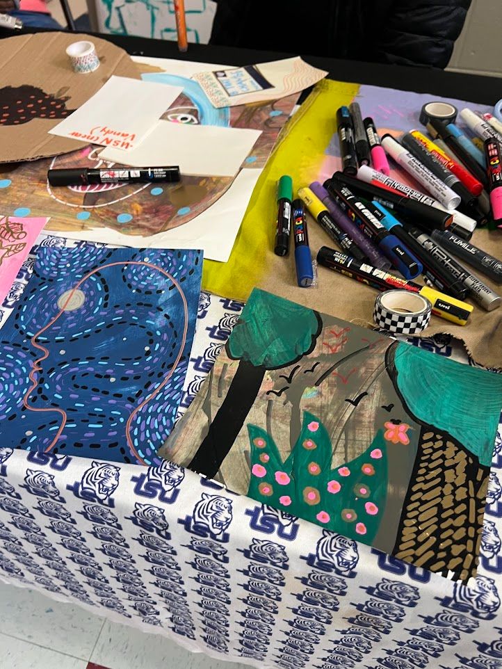 A photograph of a table with a tablecloth featuring the TSU logo that has a variety of visual art pieces and art supplies upon it.