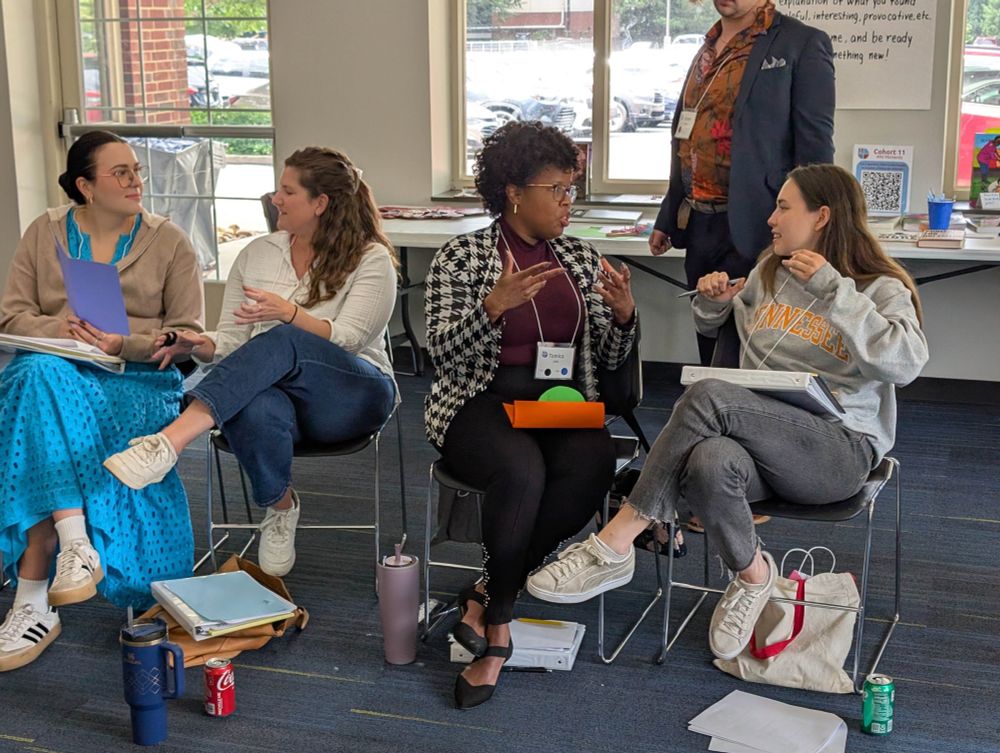 A photograph of ~4 seated educators conversing with an additional standing educator in the background in a classroom setting.