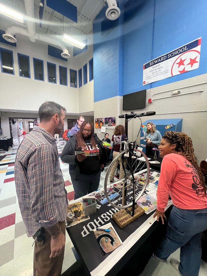 A photograph of ~7 standing educators browsing or working information booths in a school lobby setting.