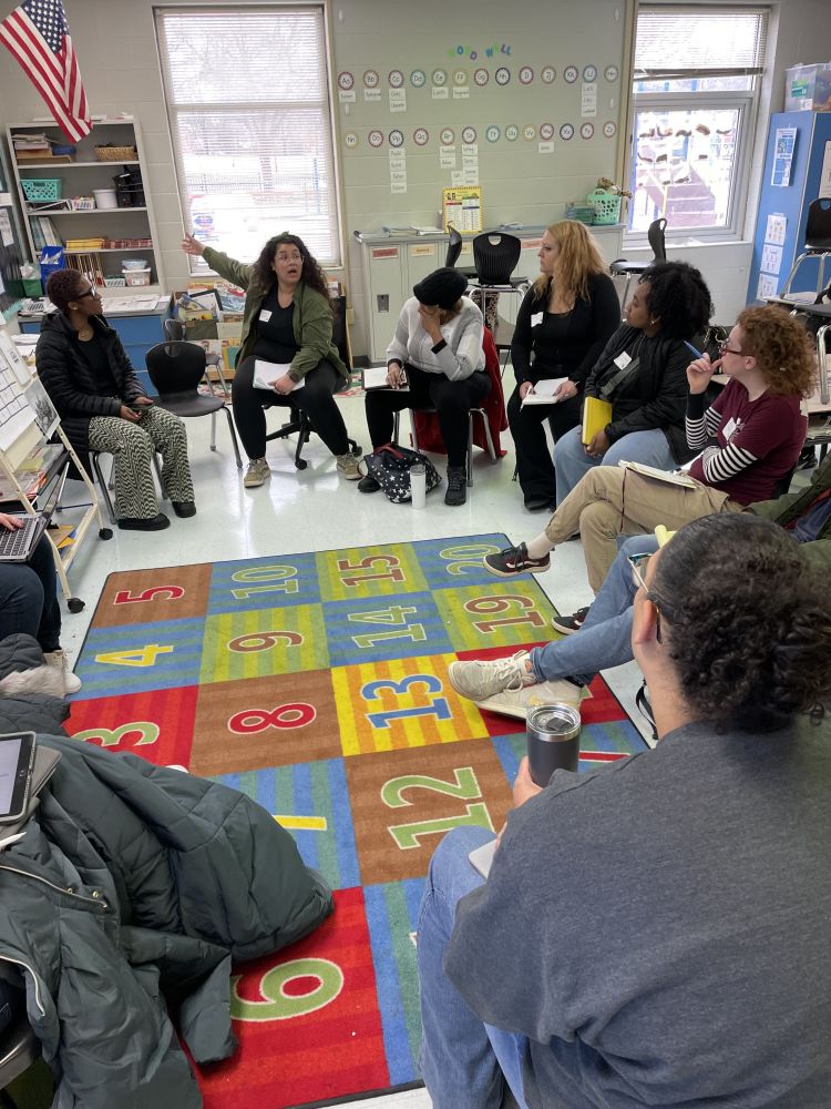 A photograph of ~9 seated educators listening to an additional seated educator who is gesturing toward an American flag speak in a classroom setting.
