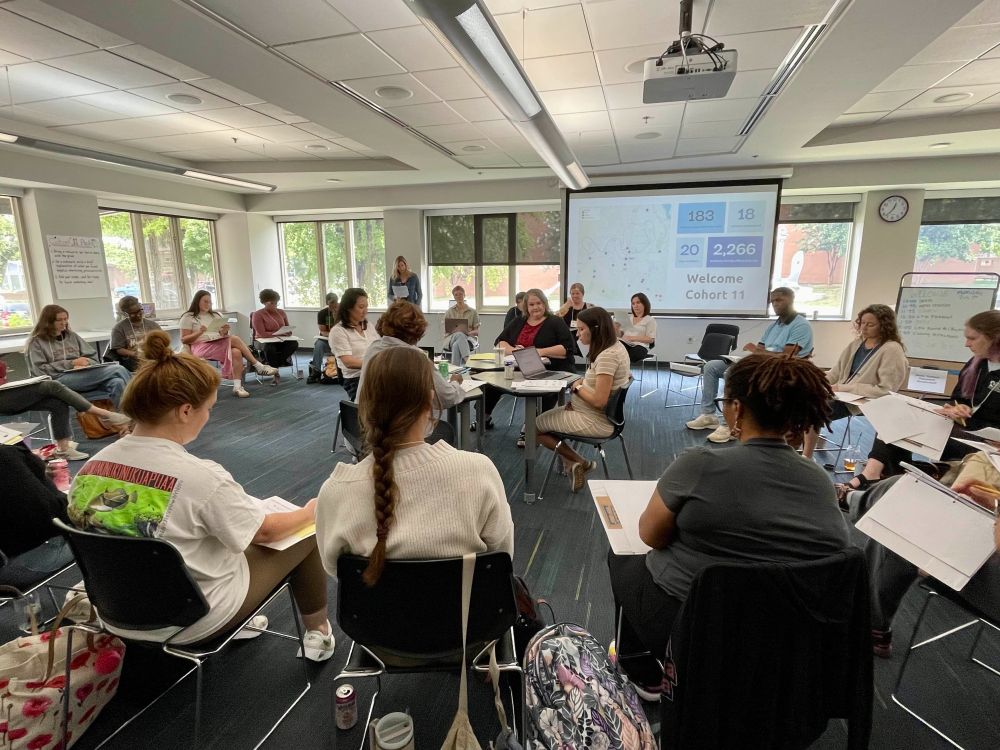 A photograph of ~23 educators in a circular formation who are engaged in a Critical Friends Group exercise in a classroom setting at EdCo Summer Workshop.