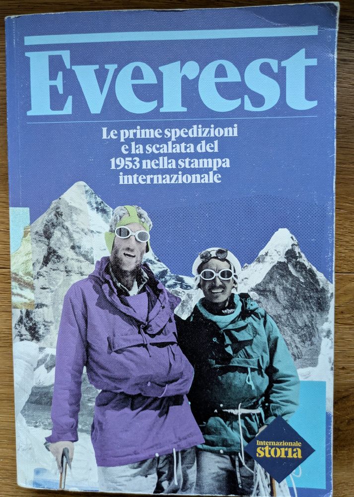 La foto mostra la copertina del libro "Everest. Le prime spedizioni e la scalata del 1953 nella stampa internazionale", pubblicato da Internazionale nel giugno 2023. 
Descrizione della copertina:
Immagine: La copertina presenta una fotografia storica in bianco e nero ritoccata, ambientata in un paesaggio montano innevato. Ritrae due alpinisti in primo piano con occhiali da ghiacciaio e abbigliamento da spedizione, sorridenti verso la macchina fotografica. L'alpinista a sinistra indossa una giacca a vento viola, mentre quello a destra ne indossa una verde scuro.
Testo: Il titolo principale, "Everest", è scritto in grandi lettere maiuscole bianche con un contorno blu che domina la parte superiore dell'immagine. Sotto il titolo, in caratteri più piccoli e bianchi, si legge "Le prime spedizioni e la scalata del 1953 nella stampa internazionale". In basso a destra è visibile il logo della collana "Internazionale storia". 
Il libro racconta la storia della prima ascensione di successo del Monte Everest, avvenuta il 29 maggio 1953 da parte di Edmund Hillary e dello sherpa Tenzing Norgay, attraverso articoli e documenti dell'epoca. 