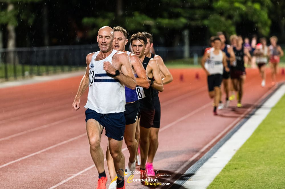 Runners on the main straight under lights at night in the rain.