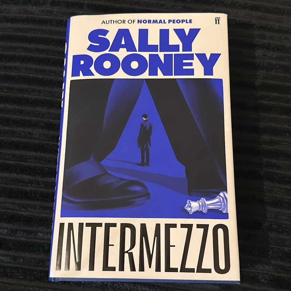 The image shows a hardcover copy of "Intermezzo" by Sally Rooney. The book cover features a striking blue and black design with "SALLY ROONEY" prominently displayed in large blue letters at the top. Below that is an artistic illustration showing a small figure of a man in a suit standing between what look like curtains or drapes, with a fallen chess piece (appears to be a white king or queen) visible to the side. The title "INTERMEZZO" is printed in large black letters at the bottom of the cover. At the very top of the cover, there's a line that reads "AUTHOR OF NORMAL PEOPLE" and the Faber & Faber publisher logo.​​​​​​​​​​​​​​​​