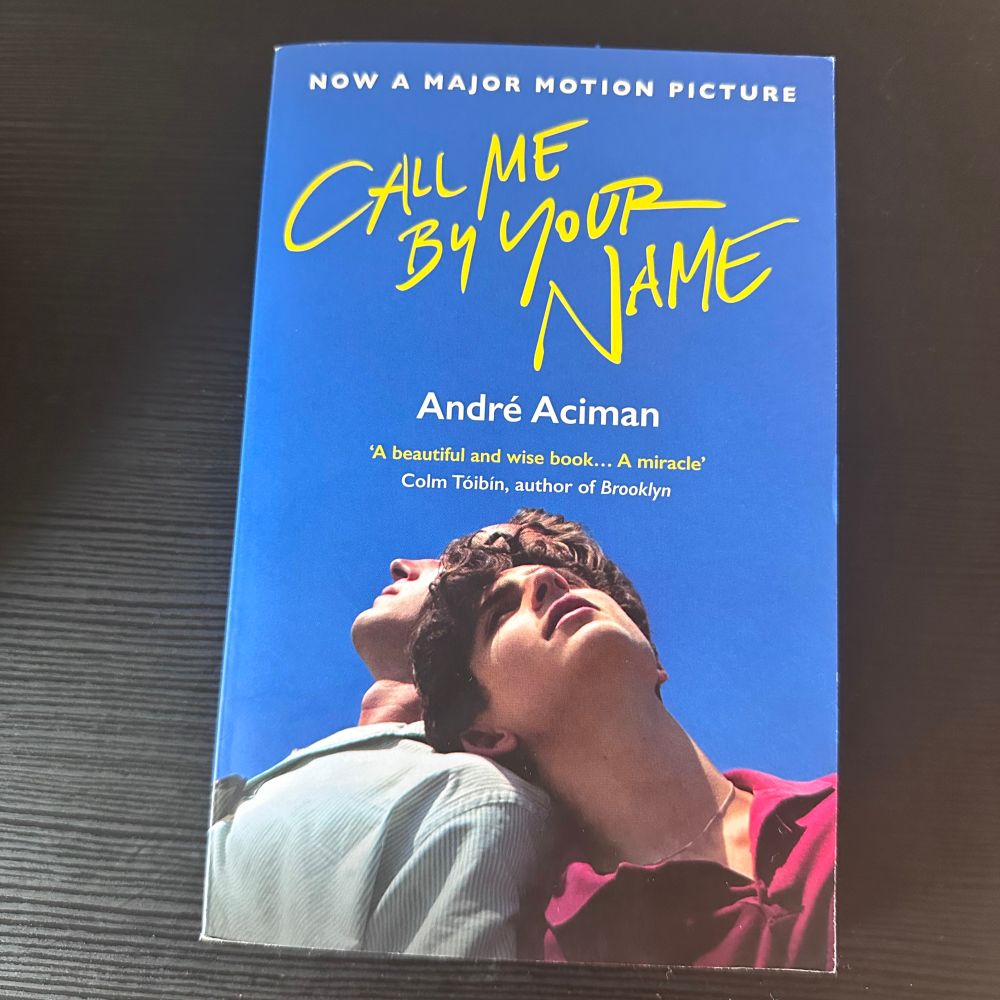 Front cover of Call Me by Your Name by André Aciman. The cover is blue with the title in large yellow handwriting-style font. It features an image of two young men leaning against each other, looking upwards against a clear blue sky. A quote from Colm Tóibín appears below the author’s name.
