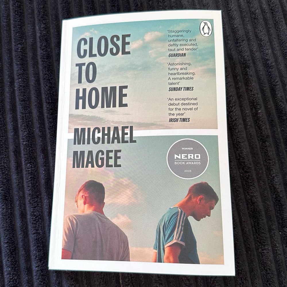 Paperback copy of the novel “Close to Home” by Michael Magee resting on a black background. The book cover features two young men looking down against a cloudy sky, with praise quotes from The Guardian, The Sunday Times, and The Irish Times. A Nero Book Awards 2023 winner sticker is also visible.