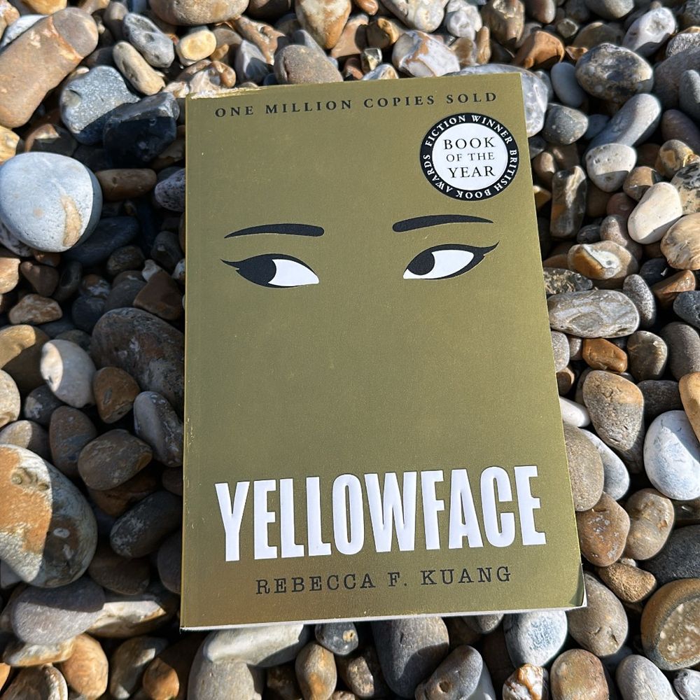 A copy of Yellowface by Rebecca F. Kuang lies on a bed of multicoloured pebbles. The book cover is gold with stylised black and white eyes near the top. A circular sticker on the cover reads “Book of the Year, British Book Awards, Fiction Winner.” The book’s edges appear slightly worn.