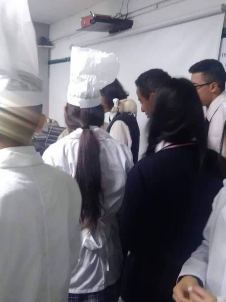 Eine Gruppe von Menschen steht in einem Raum, einige tragen weiße Kochjacken und Kochmützen. Sie scheinen sich um etwas zu versammeln, das vorne im Raum gezeigt wird. Im Hintergrund ist eine weiße Projektionsleinwand zu sehen, darüber ein Projektor. Die Atmosphäre wirkt wie in einem Kochkurs oder einer Schulung.
Eine Person hat eine Ratte mit Kochmütze auf der Schulter, wie im Film Ratatuille