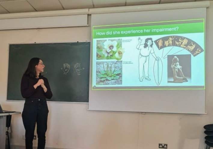 Maria stands off the the side of a pull down screen. The screen shows a slide from her presentation, showing a cartoon woman with a walking stick, an image from a greek vase, and two images of medicinal plants. Maria is standing in front of a chalkboard (which sits behind the pull-down screen) on which two hand turkeys have been drawn.