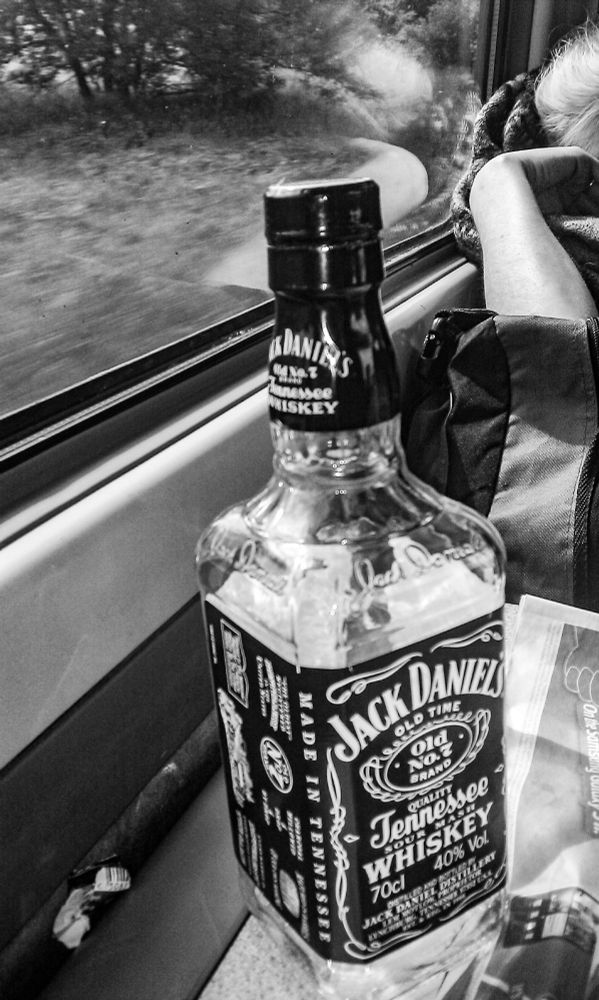 A black and white image taken on a train, the window table is shown, a persons arm, a reflection in the window, a bottle of Jack Daniels sits on the table.