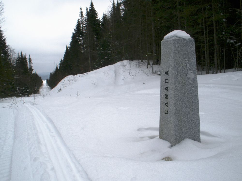 Snow covered ground with a clearing strip between the trees marking the border between Canada and US. Sled tracks with a stone border marker has Canada written on it.