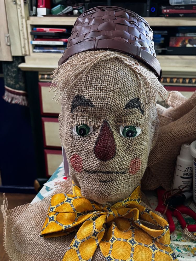 Burlap face with large green sculpted eyes looking downward, with burlap eyelids and eyelashes. The scarecrow’s neck is tied with a yellow tie with blue geometric pattern. His nose is a red teardrop shape. He has a little bit of frayed burlap hair. And he’s topped with a small brown basket as a hat.