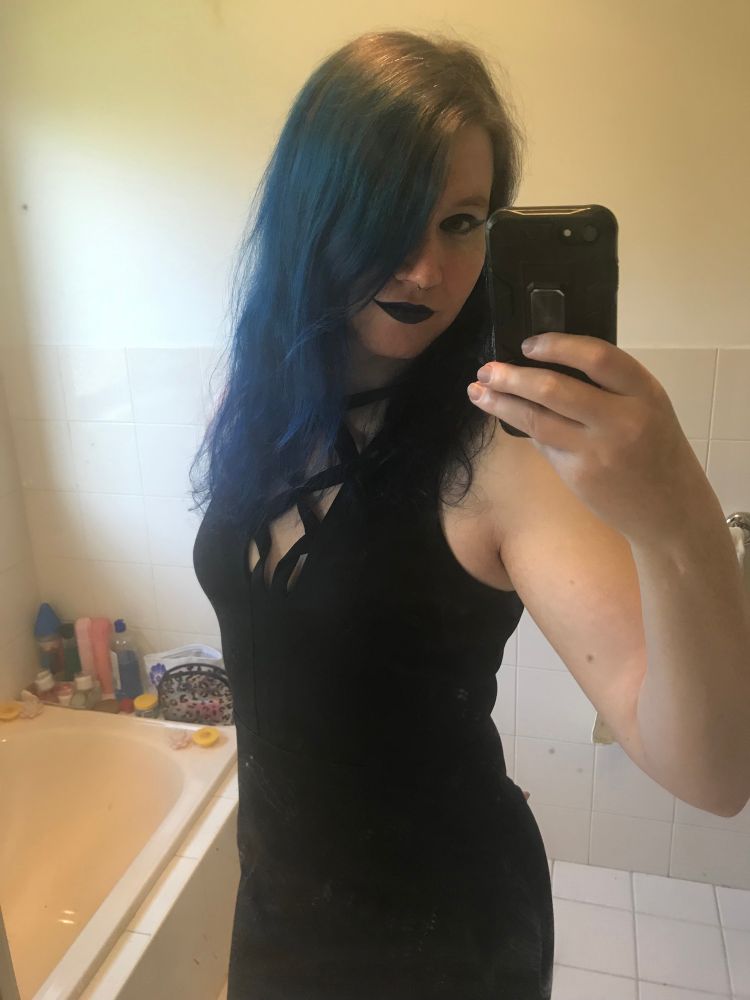 A mirror selfie of a woman in a black dress with blue hair, blue eyeshadow and black and blue lipstick